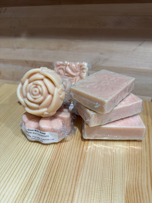Stack of handcrafted purple goat milk soap bars on wood surface, featuring rose, sunflower, and filigree shapes, scented with rose and geranium essential oils.