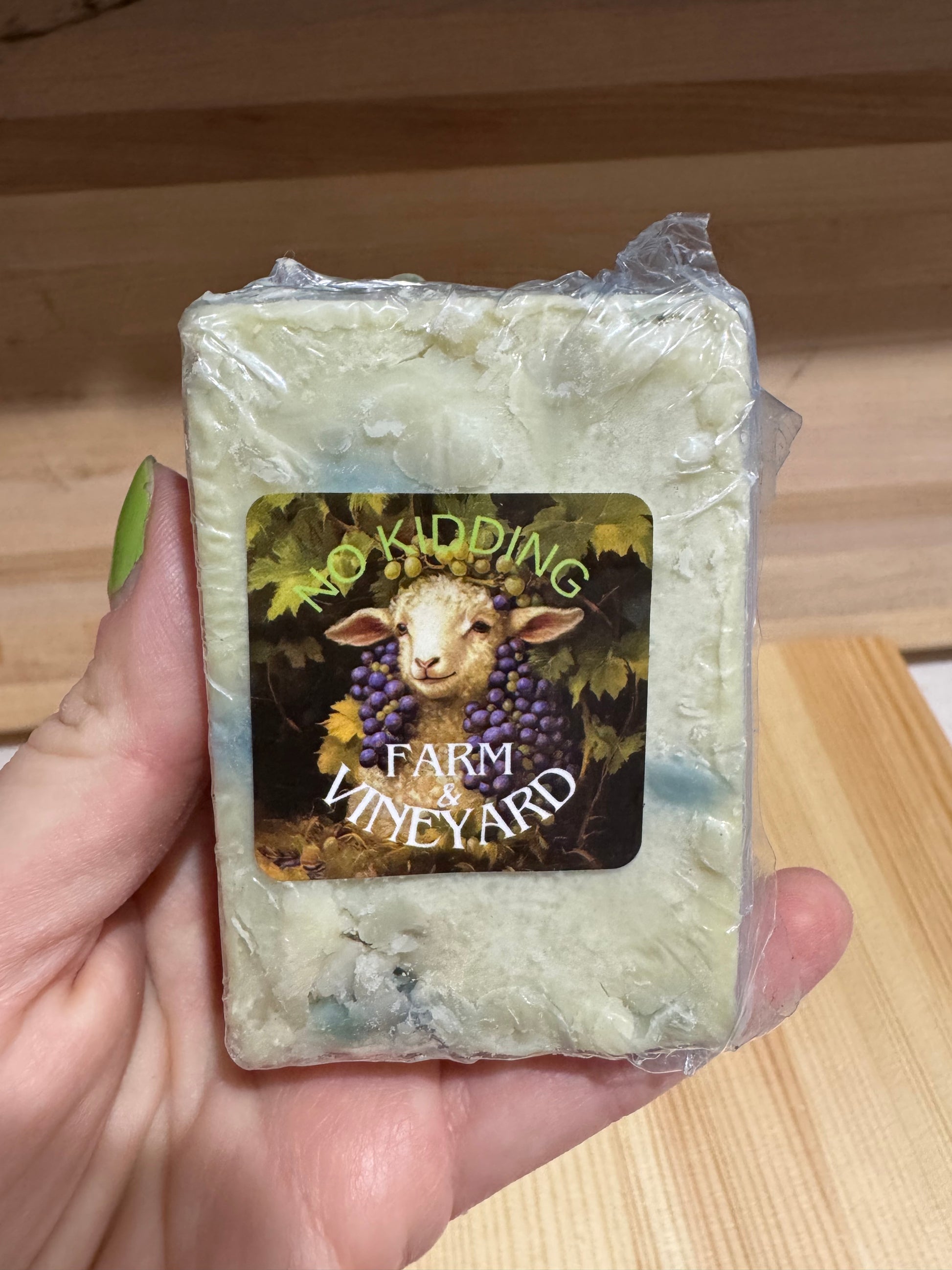 Hand holding a bar of blue and green marbled goat milk soap wrapped in shrink plastic.