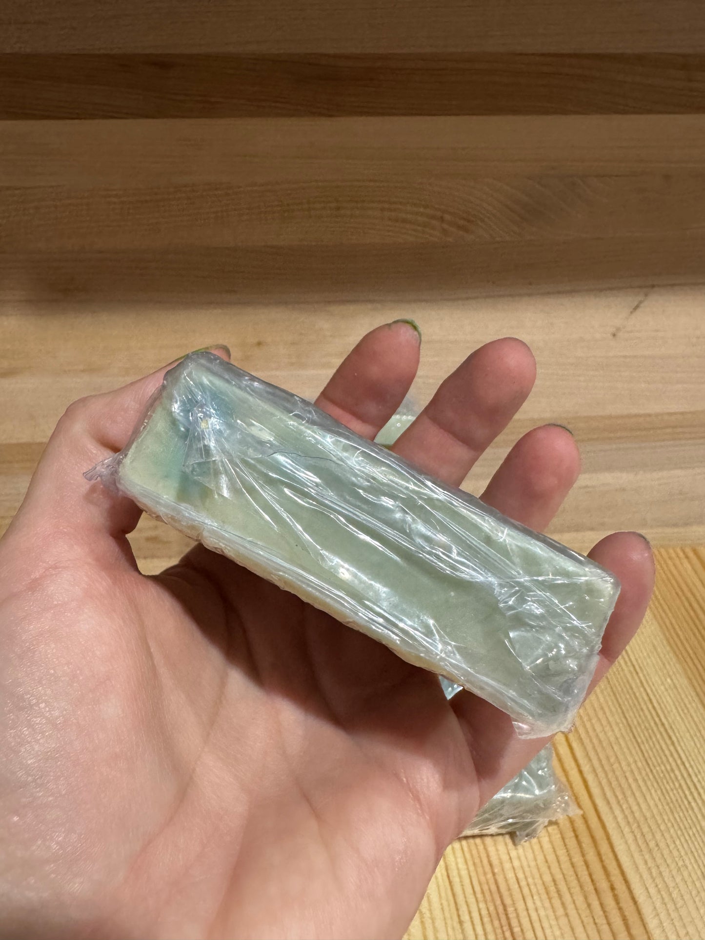 Hand holding a block of green soap wrapped in clear plastic against a wooden background